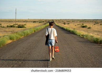 A Girl Walks Along An Empty Road In The Steppe. She Has A Ukulele In Her Hands And A Backpack On Her Shoulders. It's Summer And Warm Outside. Open Road.