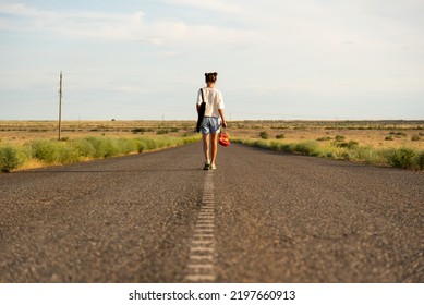 A girl walks along an empty road in the steppe. She has a ukulele in her hands and a backpack on her shoulders. It's summer and warm outside. Open road. - Powered by Shutterstock