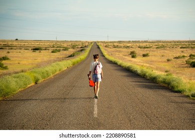 A Girl Walks Along An Empty Road In The Steppe. She Has A Ukulele In Her Hands And A Backpack On Her Shoulders. It's Summer And Warm Outside. Open Road.