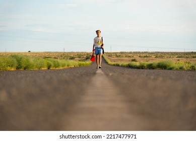 A Girl Walks Along An Empty Road In The Steppe. She Has A Ukulele In Her Hands And A Backpack On Her Shoulders. It's Summer And Warm Outside. Open Road.