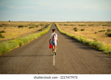 A Girl Walks Along An Empty Road In The Steppe. She Has A Ukulele In Her Hands And A Backpack On Her Shoulders. It's Summer And Warm Outside. Open Road.