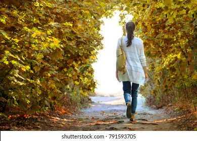 The girl walks along the path in the autumn woods to the light in a white jacket and jeans.