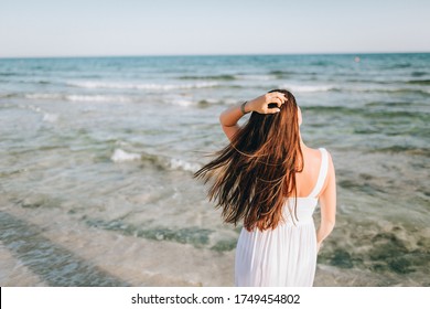 Girl Walks Along The Sea. Back View. A Woman Alone On The Sea Shore Admires The Evening Landscape