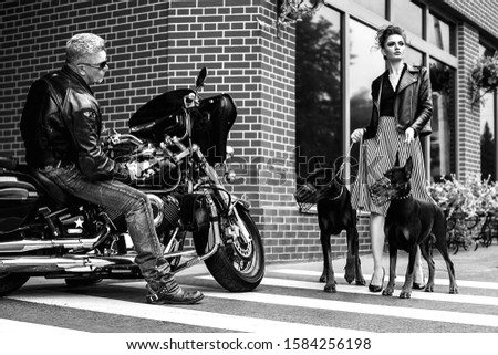 A girl walks along the street in the city along the building with two Dobermans on a leash. A man on a motorcycle looks at the girl as she crosses the road with dogs and she crosses the road with dogs