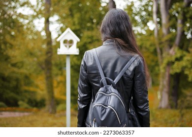girl walks in an spring park, young lady back view. woman in black jacket with backpack. natural green background, bokeh, focus. wooden birdhouse. white bird feeder and girl in the garden, autumn