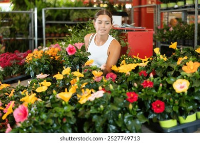 Girl walks through exhibition of ornamental plants, examines hibiscus in showcase. Visitors to wholesale flower market stand against background of shopping malls with decorative flowering plants - Powered by Shutterstock