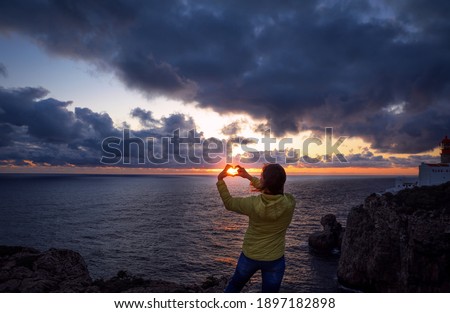 Girl watching the beautiful sunset at Cape of St. Vincent in Portugal. Storm clouds over the Atlantic ocean during sunset. The girl hold her hans in the shape of a heart, in the center of which is sun
