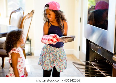 Girl Watching Big Sister Insert Cake Tin Into Oven, Face To Face Smiling