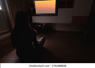 Girl Watching Tv In A Dark Room. Back View. Woman Sitiing On Floor In Front Of Television Set. Watch Movies And Shows In A Comfortable Home Environment