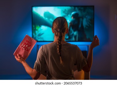 The girl is watching TV. She has a pack of popcorn in her hand. Cozy home environment, relaxation, rest, quarantine, self-isolation. Shooting from the back. - Shutterstock ID 2065537073