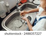 Girl, water and learning to wash dishes in kitchen, sanitary and prevent bacteria or germs. Female person, child and soap for cleaning mess in home, housework and kid helping for responsibility