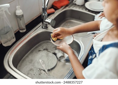 Girl, water and learning to wash dishes in kitchen, sanitary and prevent bacteria or germs. Female person, child and soap for cleaning mess in home, housework and kid helping for responsibility - Powered by Shutterstock