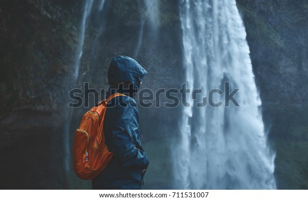 Girl\
in waterproof clothing stands under the Seljalandsfoss waterfall in\
Iceland. back view, woman with small orange\
backpack
