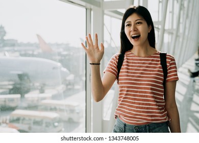 Girl Waves Her Hand On Arrival Lounge At Airport On Summer Vacation Travel Back Home. Friends Pick Her Up Cheerfully Saying Hi Walking In Hall Near Window With Airlines In Background On Runways.