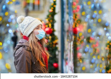 Girl wearing face mask on a Parisian street or at Christmas market looking at shop windows decorated for Christmas. Seasonal holidays during pandemic and coronavirus outbreak