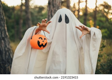 Girl wearing ghost costume holding pumpkin bucket with candies, standing in a forest on Halloween. - Powered by Shutterstock