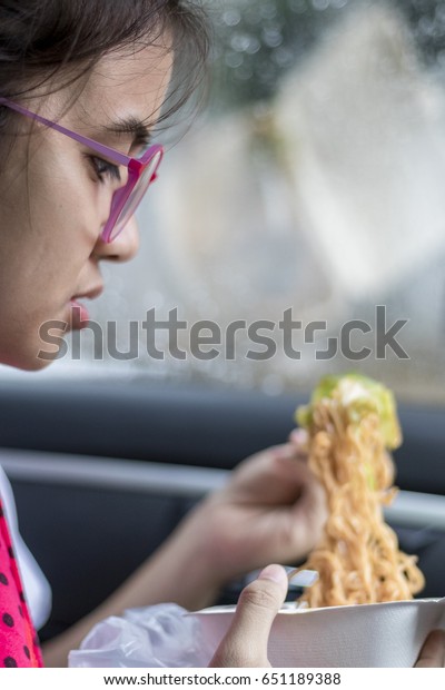 \
The girl\
wearing glasses sitting down to eat noodles in the car between the\
seams on a signal.A young girl wearing glasses sitting on the bear\
in the car during the traffic\
jam.