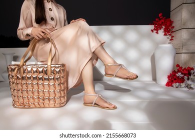 The girl is wearing a golden dress, has golden black buttons on the dress, and is wearing golden sandals, is wearing a golden purse in her hand, and is sitting on the bench, with flowers in front. - Shutterstock ID 2155668073