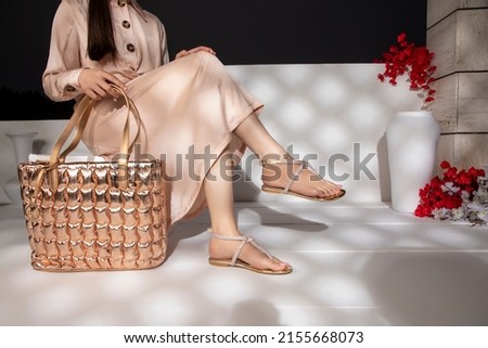 The girl is wearing a golden dress, has golden black buttons on the dress, and is wearing golden sandals, is wearing a golden purse in her hand, and is sitting on the bench, with flowers in front.