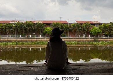 A Girl Wearing A Hat, Sitting At The Wooden Dock Near The Canal, Looking The Water Far Away. The Opposite Site Is A Two Stories Timber Building And A Line Of Betel Palm Tree.
