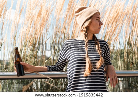 Girl wearing hat and t-shirt posing with bottle of cold-brew coffee outdoor in front of reeds in the center of European city