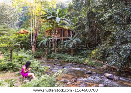 A girl wearing a hill tribe costume sitting use smartphone by the stream In a relaxing forest scene.