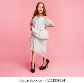 Girl Wearing Large Dress And High Heels Shoes Pretending To Be A Grown Up Woman. Girl Child Posing In Oversized Clothes On Pink Background.