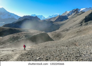 A girl wearing pink jacket hikes up through the dust storm. She is holding her hands close to the body, not to lose temperature. Clear sky. High altitude Himalayan trek. Harsh and barren landscape.
