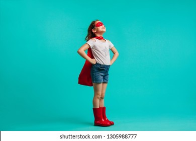 Girl wearing red gumboots, cape and eye mask standing with her hands on hips in studio. Super girl looking away on blue background.