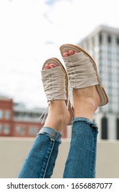 Girl Wearing White Leather Sandals Kicking Feet Up With City Buildings In Background