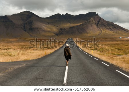 Girl wearing a woolly hat and wellington boots walks in the center of a quiet road towards a huge volcanic mountain in the distance, near Snaefellsjokull national park, Iceland.