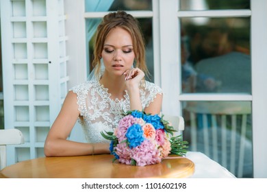 The Girl In The Wedding Dress Sits At The Table Alone And Looks Upset. The Bride Looks Sadly At A Bouquet Of Flowers.