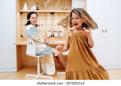 Girl Whirls In The Children's Room Against The Background Of Her Mother Sitting At The Table. A Real Life Moment