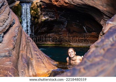 A girl in a white bikini relaxes in a rock pool with a waterfall cascading over her head; swimming in natural pools in karijini national park, western australia