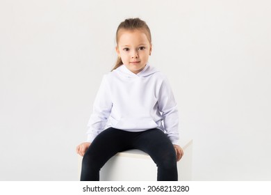 Girl In A White Blank Hoodie Sits On A White Background. Kids Hoodies Mockup.