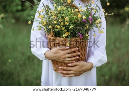 A girl in a white blouse holds a wicker basket with a bouquet of wild flowers. Summer walk in the field. midsection.