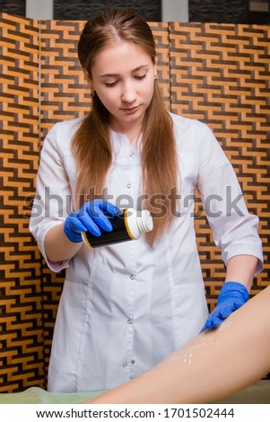 A girl in a white coat, a depilation master at work in a beauty salon. The specialist removes the hair on the legs of the client.