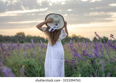 girl in a white dress and hat in a lupine field looks at the sunset