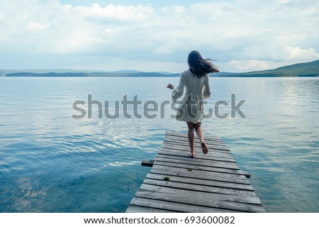 A girl in a white dress is running along a wooden pier. On a background of blue mountains with water (ocean, sea, lake) and clouds. Fun, happiness concept