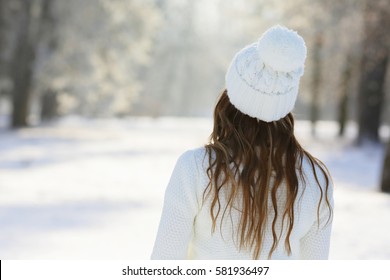 Girl in a white hat with a pompon, standing back in the winter park