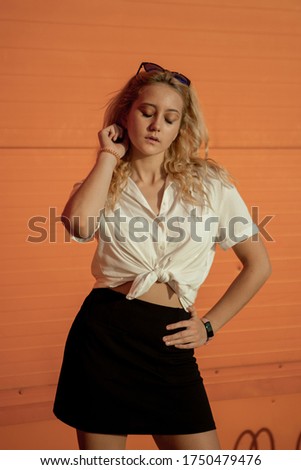 A girl in a white shirt against an orange wall. Blonde in sunglasses.