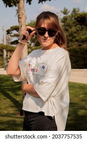 Girl In A White T-shirt And Black Glasses Portrait Photography Against The Background Of Green Trees In The Park In Warm Shades