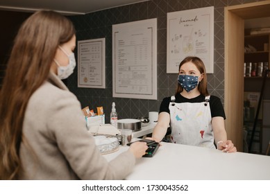 A Girl Who Wears A Medical Face Mask Uses A Smartphone To Pay By NFC Technology. A Female Barista In A Face Mask Holds Out A Terminal For Contactless Paying To A Client.