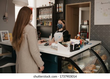 A Girl Who Wears A Medical Face Mask Uses A Smartphone To Pay By NFC Technology. A Female Barista In A Face Mask Holds Out A Terminal For Contactless Paying To A Client.