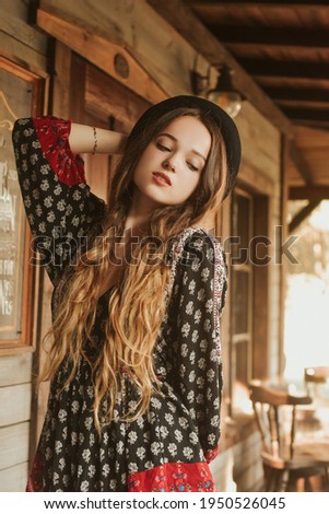 Girl in the Wild West, in Western house. Girl in hat with long cerly hair smile. Beautiful pretty girl in black hat. Incredible trip, travel. Woman on the terrace in red dress pouse.