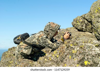 Girl Wildlife Photographer In Camouflage Suit In The Mountains Hiding Behind A Stone Photographing Something