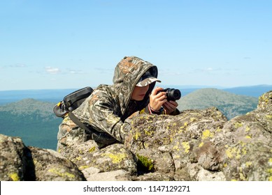 Girl Wildlife Photographer In Camouflage Suit In The Mountains Hiding Behind A Stone Photographing Something