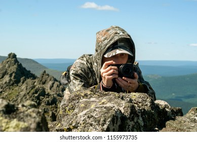 Girl Wildlife Photographer In Camouflage Suit In The Mountains Hiding Behind A Stone Photographing Something