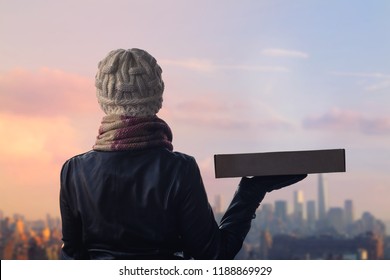 A Girl In Winter Clothes With A Box For Pizza On The Manhattan Background. New York City.   