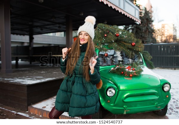 Girl in winter green clothes on\
the street, near a green retro car. Beautiful happy woman with gift\
box at celebration party. Christmas and New Year\
holidays.